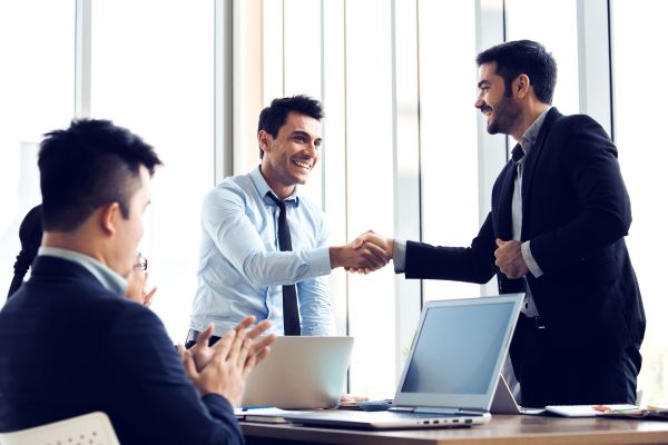 Business professionals shaking hands in a bright conference room while a colleague observes.